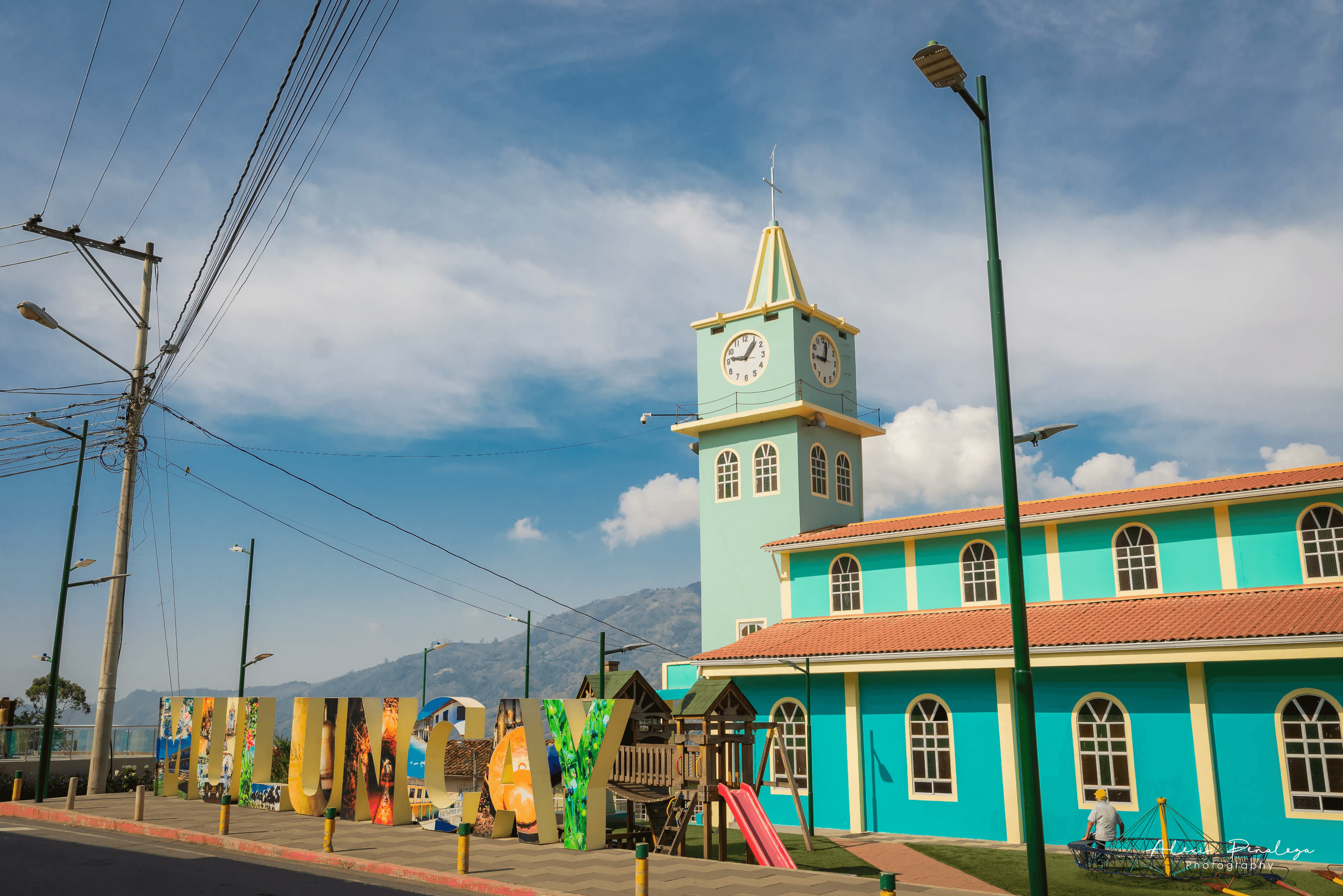 Letrero de Muluncay junto a juegos infantiles con la torre de la iglesia de fondo